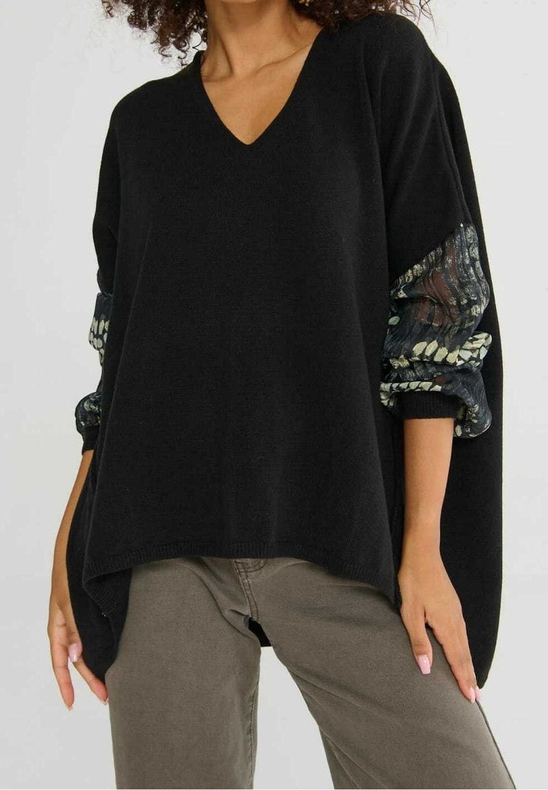 Femme portant un pull ample noir à col en V avec des manches transparentes à motifs, associée à un pantalon vert olive, debout avec les mains détendues.