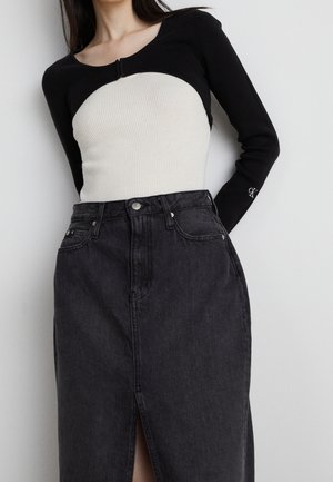 Femme portant un cardigan noir court par-dessus un haut blanc côtelé et une jupe en denim noire taille haute avec une fente à l'avant.