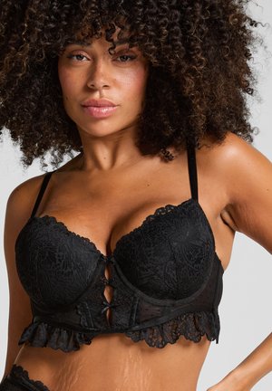 Donna con capelli ricci che indossa un bralette di pizzo nero con motivi floreali e bordi arricciati, posando contro uno sfondo neutro.