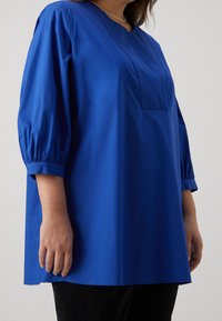 Blå, luftigt passande topp med 3/4 puffärmar och fyrkantig halsringning. Har en bröstfick och ett mjukt, lätt tyg.