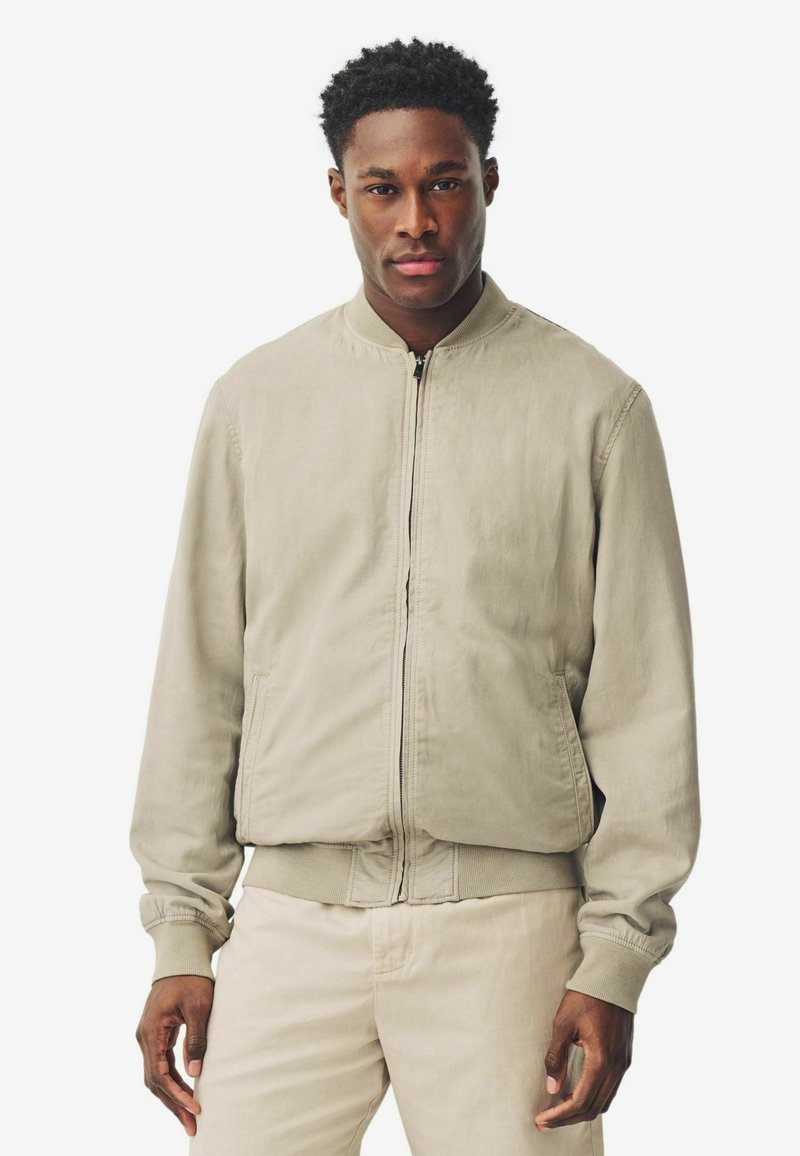 Giacca bomber leggera beige con chiusura frontale a zip, polsini a costine e design a girocollo. Tessuto liscio con tasche laterali.