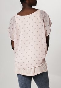 Blusa de manga corta rosa claro con tela transparente, presenta lunares grises a rayas, escote redondeado y dobladillo en capas.
