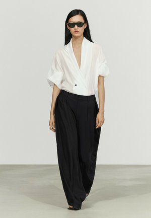 Donna con occhiali da sole neri, camicetta bianca larga con maniche arrotolate e pantaloni neri larghi che cammina su uno sfondo neutro di passerella.