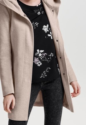 Abrigo de lana beige con capucha, botones delanteros y bolsillos laterales, superpuesto sobre una blusa negra con estampado floral y jeans oscuros.