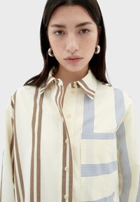 Beige Button-up-Hemd mit braunen und hellblauen vertikalen Streifen; verfügt über eine aufgesetzte Tasche und einen klassischen Kragen. Glatte Baumwolltextur.
