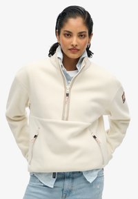 Pullover in pile off-white con mezza zip, due tasche frontali e accentuazioni ricamate. Indossato sopra una camicia a righe blu.