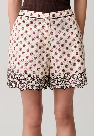 Pantalones cortos color crema con pequeño estampado floral rojo y ribete floral negro con bordes ondulados en el dobladillo, usados con una camiseta oscura de canalé.