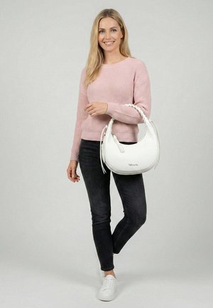 Donna sorridente, indossa un maglione rosa e jeans neri, tiene in mano una borsa bianca con manico intrecciato, in piedi davanti a uno sfondo semplice.