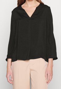 Kvinna som bär en lös svart långärmad blus med V-hals och beige högmidjade byxor, med avslappnade händer vid sidorna.