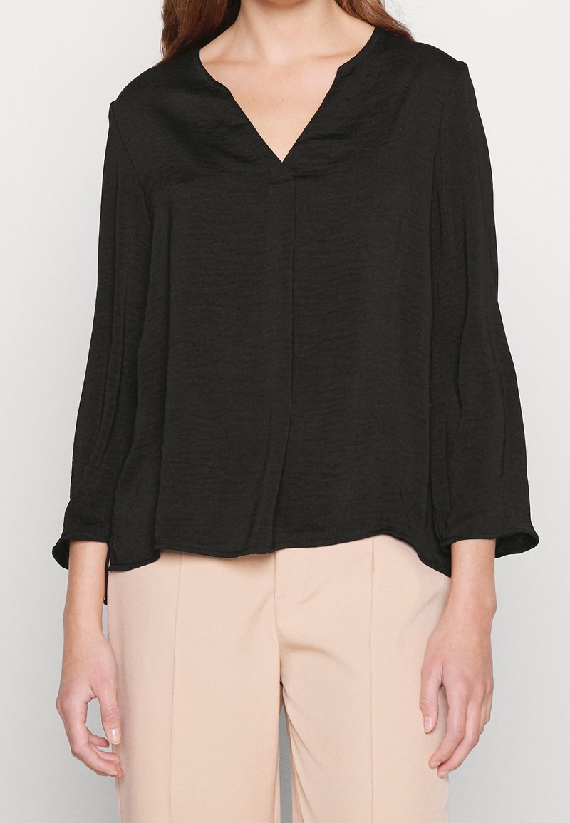 Kvinna som bär en lös svart långärmad blus med V-hals och beige högmidjade byxor, med avslappnade händer vid sidorna.