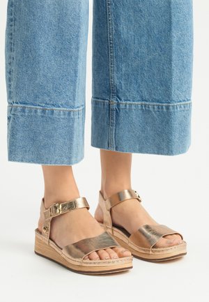 Sandales à talon compensé dorées métalliques avec une large bride au niveau des orteils, une boucle à la cheville et une semelle en bois texturée, portées avec un jean skinny bleu cropped.