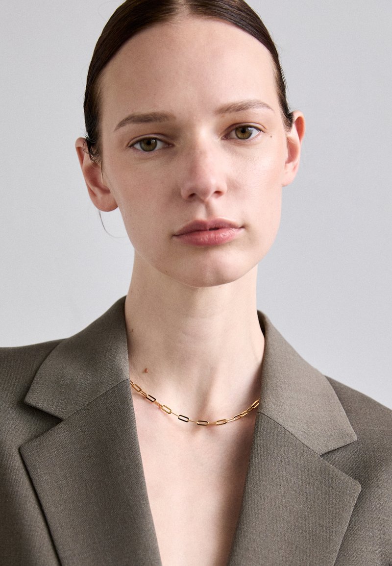 Kuldne kett, mis koosneb vahelduvatest piklikest ja ovaalsetest linkidest, kantud halli bleiseri peal, puhta kaelani ja minimaalsete tekstuuridega.