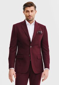 Bordowy garnitur z wyraźnie zbudowanymi klapami, dwoma przednimi kieszeniami i slim fit. Połączony z białą koszulą i chusteczką w kropki.