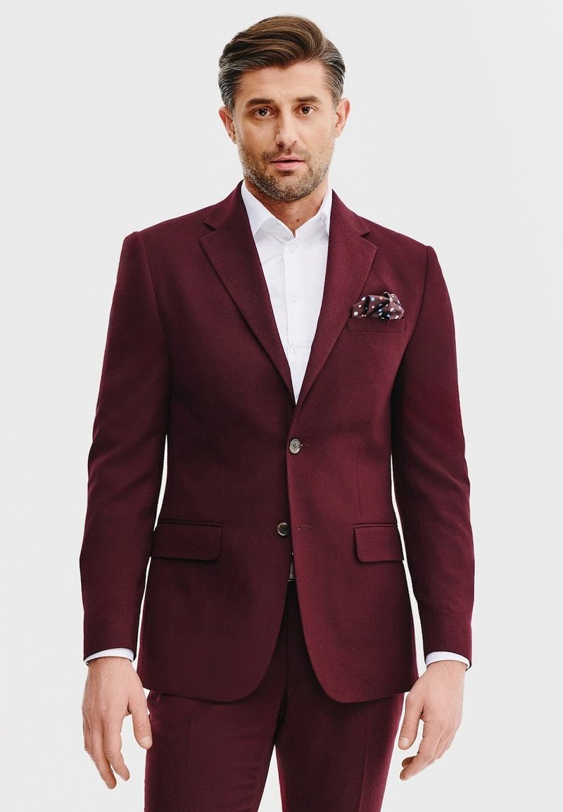 Bordowy garnitur z wyraźnie zbudowanymi klapami, dwoma przednimi kieszeniami i slim fit. Połączony z białą koszulą i chusteczką w kropki.