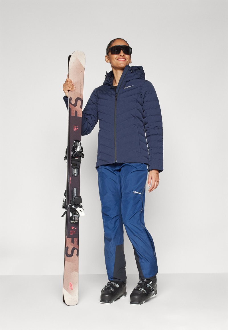 Marineblaues Pufferjacke mit Kapuze, blaue Skihosen und schwarze Skistiefel. Hält ein Paar Skier mit Bindungen, pinke und braune Skifläche.