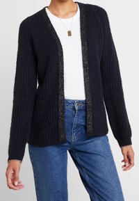 Cardigan lavorato a maglia di colore blu navy scuro con motivo a trama, apertura frontale e accenti di filo metallico lungo i bordi. Presenta maniche lunghe.