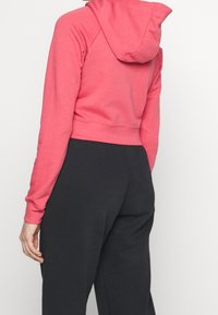 Krátká mikina s kapucí v jasně růžové barvě, s kapucí a dlouhými rukávy, vyrobená z měkké látky; doplněná černými kalhotami s volným střihem.
