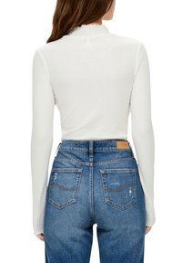 Bílý žebrovaný top s dlouhými rukávy a vlnitým výstřihem, doplněný o vysoké modré džíny s jemným poškozením a nášivkou.