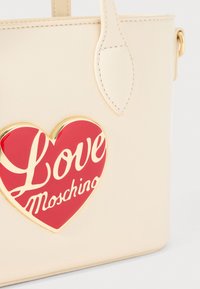 Beige handbag made of smooth material, featuring a prominent red heart-shaped logo with "Love Moschino" in white script. Gold-tone accents.