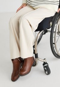 Brune lær ankle boots med rundt tå og teksturert såle, båret med kremfargede vide bukser, en stripete genser, og en rullestol synlig.