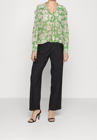 Groene bloemenblouse met lange mouwen, geplooide textuur en een strikdetail bij de hals, gecombineerd met zwarte wijde broek en sandalen met banden.