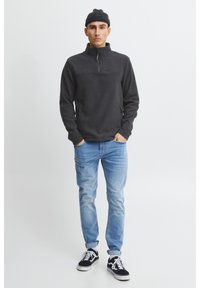 Temno siva flis pulover s polovičnim zadrgo in ovratnikom, oblečen s svetlo modrimi jeans hlačami in črnimi športnimi copati, z dvema stranskima žepoma.