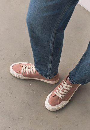 Person iført blå jeans og lyserøde lærredsneakers med hvide snørebånd, stående på en tekstureret grå gulv.