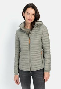 Veste légère verte padée avec une capuche, fermeture éclair à l'avant, poches latérales avec des accents beige, et poignets côtelés. Portée sur un haut gris.