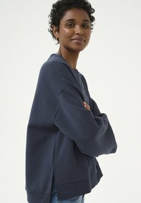 Tumši zila pārāk liela sporta krekla, izgatavota no mīkstas auduma, ar pazeminātām plecu līnijām un sānu šķeltnēm, kas nodrošina kustību brīvību.