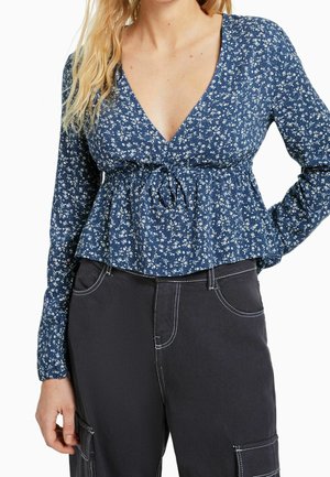 Blusa a fantasia floreale blu con maniche lunghe, scollatura a V profonda e vita arricciata. Abbinate a pantaloni scuri con dettagli di cucitura bianca.
