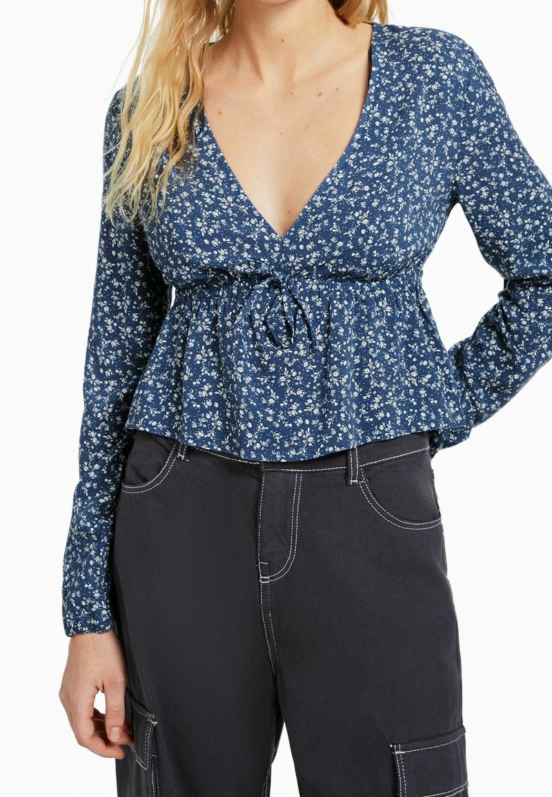 Blusa a fantasia floreale blu con maniche lunghe, scollatura a V profonda e vita arricciata. Abbinate a pantaloni scuri con dettagli di cucitura bianca.