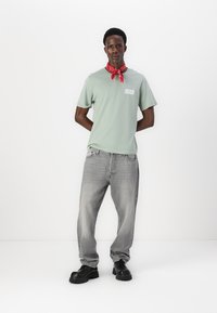 Lys grønn T-skjorte med hvit logo, grå jeans og svarte klumpete sko. Et rødt bandana er knyttet rundt halsen. Glatt stoff, uformell passform.