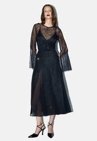 Top in pizzo nero con maniche a campana trasparenti, sovrapposto a una gonna scura e luccicante con fessure frontali e una finitura liscia. Abbinato a tacchi neri.