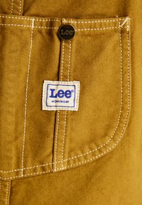 Brūns audums ar tekstūru, kurā ir balta birka ar zilu tekstu "Lee WORKWEAR" un zeltīta pogas aizdare kabatā.