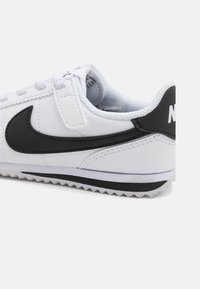 Bijela sintetička tenisica s crnim Nike logom, teksturiranim gornjim dijelom, zatvaranjem na vezivanje i gumenim potplatom s uzorkom za prianjanje.