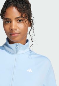 Casaco azul claro com fecho éclair, com um colarinho alto, apresentando detalhes do logótipo brancos e uma textura de tecido suave e leve.