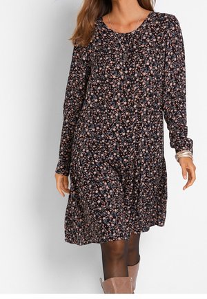 Femme portant une robe noire à manches longues avec un petit imprimé floral rose et bleu, accessoirisée avec des colliers, des bracelets, des collants noirs et des bottines marron clair.