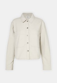 Chaqueta cortada de color beige claro hecha de algodón suave, con un cuello clásico, cierre de botones y dos bolsillos delanteros de parche.