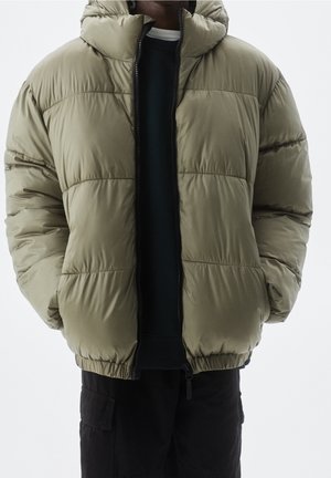 Personne portant une doudoune vert olive sur un t-shirt noir et un pantalon cargo noir, debout les mains dans les poches de la veste.