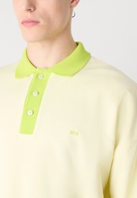 Camisa polo de color amarillo claro con un cuello verde en contraste. Presenta tres botones y un sutil bordado "MN" en verde. Textura suave.