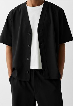 Osoba nosząca czarną koszulę z krótkim rękawem na białym t-shircie, z rękami w kieszeniach czarnych spodni, na jednolitym tle.