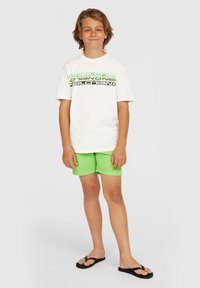 Witte t-shirt met grote zwarte en neon groene tekstpatronen. Heldere groene shorts en zwarte flip-flops maken de outfit compleet.