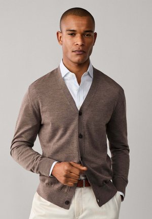 Homme portant un cardigan marron boutonné sur une chemise blanche à col, pantalon crème et ceinture marron, debout devant un fond uni.