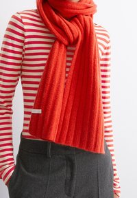 Personne portant une chemise à manches longues rayée rouge et blanche, un pantalon gris foncé, et une écharpe rouge épaisse à côtes enroulée autour du cou.