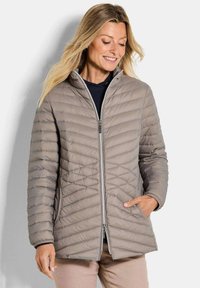 Vrouw in een lichtbruine gewatteerde jas met een ritssluiting aan de voorkant en een hand in de zak, glimlachend en naar beneden kijkend met los blonde haar.