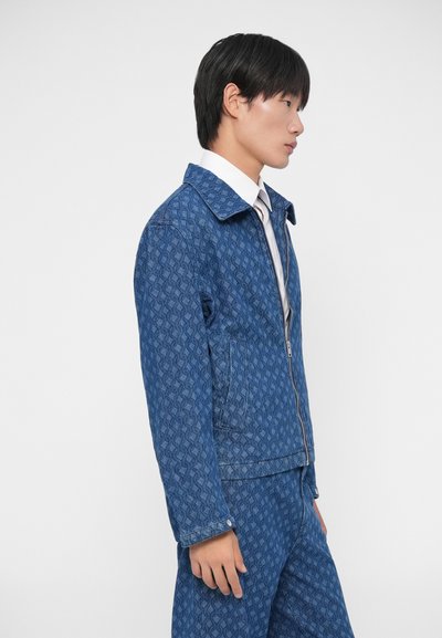 Chaqueta de mezclilla azul con un patr�ón geométrico, cierre frontal y un cuello clásico. El modelo lleva una camisa blanca debajo.