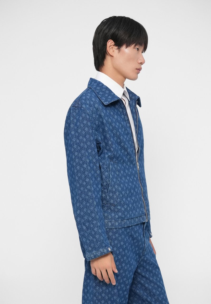 Blue denim jacket featuring a geometric pattern, front zipper, and a classic collar. Model is wearing a white shirt underneath.