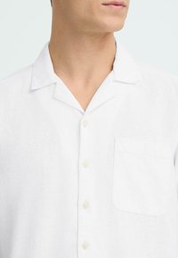 Chemise blanche en lin à manches courtes avec un col classique, une fermeture à six boutons et une poche poitrine unique en tissu texturé.