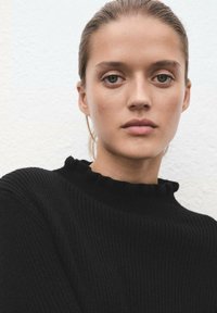 Maglione nero a costine con un collo alto e arricciato. Texture liscia e design aderente. La modella ha un'espressione neutra e un trucco leggero e naturale.