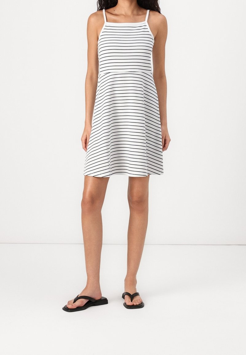 Femme portant une robe blanche sans manches longueur genou avec de fines rayures horizontales noires et des sandales plates noires, debout sur un fond blanc.